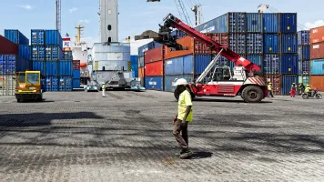 A black man in a white baseball cap walks in front of colorful shipping containers at a loading dock.