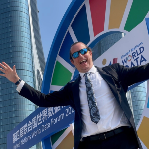 A man in a suit and sunglasses smiling with his arms out in front of a brightly colored sign.