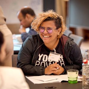 Jimena Cascante Matamoros sitting at a table and smiling, opposite two other people. She has an open notebook in front of her, and wears a t-shirt that says 'youth power'