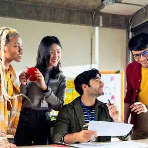 Four young people working together around a desk, one is sitting and holding a piece of paper, the other three are standing around the desk