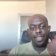 A man on a video speaking to the camera in a yellow shirt with his left hand raised in expression.