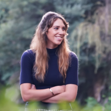 Person in a blue short with long, blonde hair standing in front of trees with arms crossed and smiling.