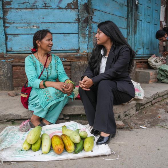 Two people seated on a sidewalk curb, facing each other, in front of a pile of fruit on the ground.
