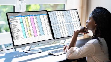 A black woman sits in front of dual monitors with colorful text in front of a window. It is daylight outside. Her back is toward the camera.