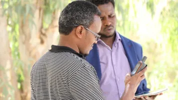 Two men standing in front of trees looking at mobile phones.