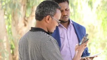 Two men in front of trees look at mobile phones.