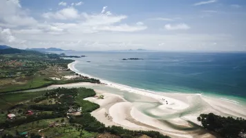 An aerial view of a green coast line and the sea with a white sand beach and clouds in the background.