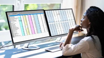 A black woman sits in front of dual monitors with colorful text in front of a window. It is daylight outside. Her back is toward the camera.