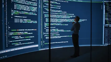 man standing in front of a wall-length computer screen, looking at coding data