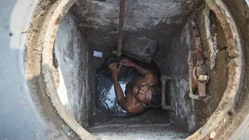 A man with dark skin and a moustach stares up from a sewage hole. 