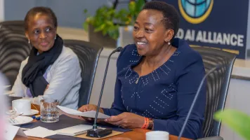 A black woman seated at a table with short hair and a blues shirt smiles while speaking into a microphone. A black woman in a gray shirt and black scarf to her right at the table smiles at her.