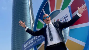 A man in a suit and sunglasses smiling with his arms out in front of a brightly colored sign.