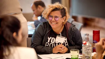 Jimena Cascante Matamoros sitting at a table and smiling, opposite two other people. She has an open notebook in front of her, and wears a t-shirt that says 'youth power'