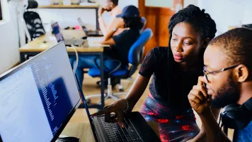 Two people sitting in front of a desktop computer and working together. One person is pointing at the screen. Photo by Desola Lanre Ologun, via Unsplash