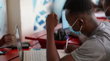 Person in gray t-shirt sitting in front of laptop, wearing earbuds. Photo by Kojo Kwarteng, via Unsplash
