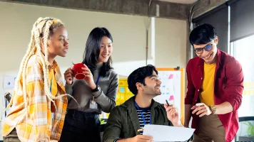 Four young people working together around a desk, one is sitting and holding a piece of paper, the other three are standing around the desk