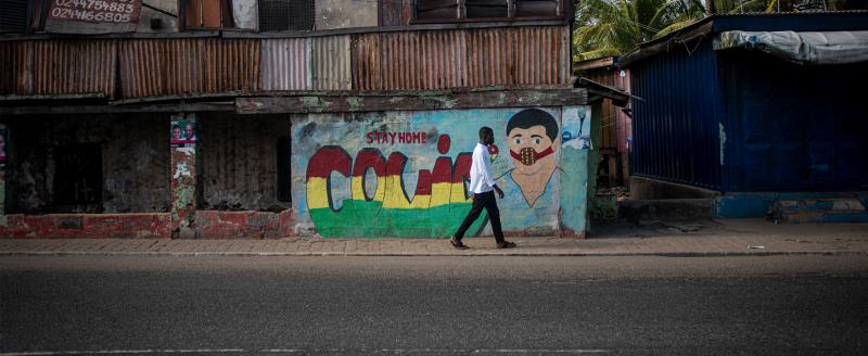 Man walking across an empty street in Accra, Ghana, with "COVID-19" graffitied on the wall