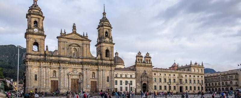 A stone church with two towers in the background of a plaza with people and birds in the foreground.
