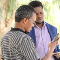 Two men in front of trees look at mobile phones.