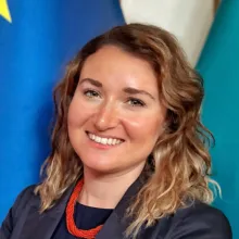 A young, white woman with long blonde hair wearing a blue suit and smiling at the camera in front of a blue flag with yellow stars.