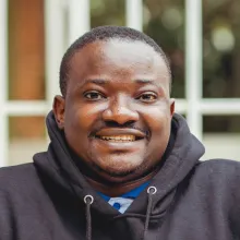 A black man with short hair smiling at the camera wearing a black hoodie.