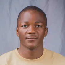 A young, black man with very short hair in a yellow t-shirt facing the camera and smiling slightly.