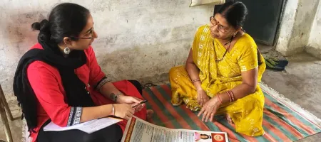 Two women seated on the floor facing each other wearing colorful saris. A large, colorful page is on the floor between them.