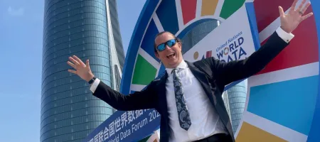 A man in a suit and sunglasses smiling with his arms out in front of a brightly colored sign.