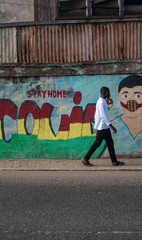Man walking across an empty street in Accra, Ghana, with "COVID-19" graffitied on the wall