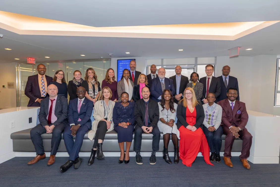 Two rows of people in a group photo. The front row is seated on a white couch. The crowd is diverse in terms of age, race, and gender, and people are wearing business clothes.